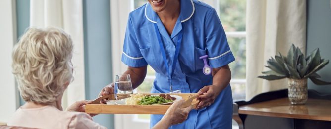 female-care-worker-in-uniform-bringing-meal-on-tray-to-senior-woman-sitting-in-lounge-at-home.jpg