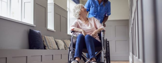 senior-woman-at-home-being-pushed-in-wheelchair-by-female-care-worker-in-uniform.jpg